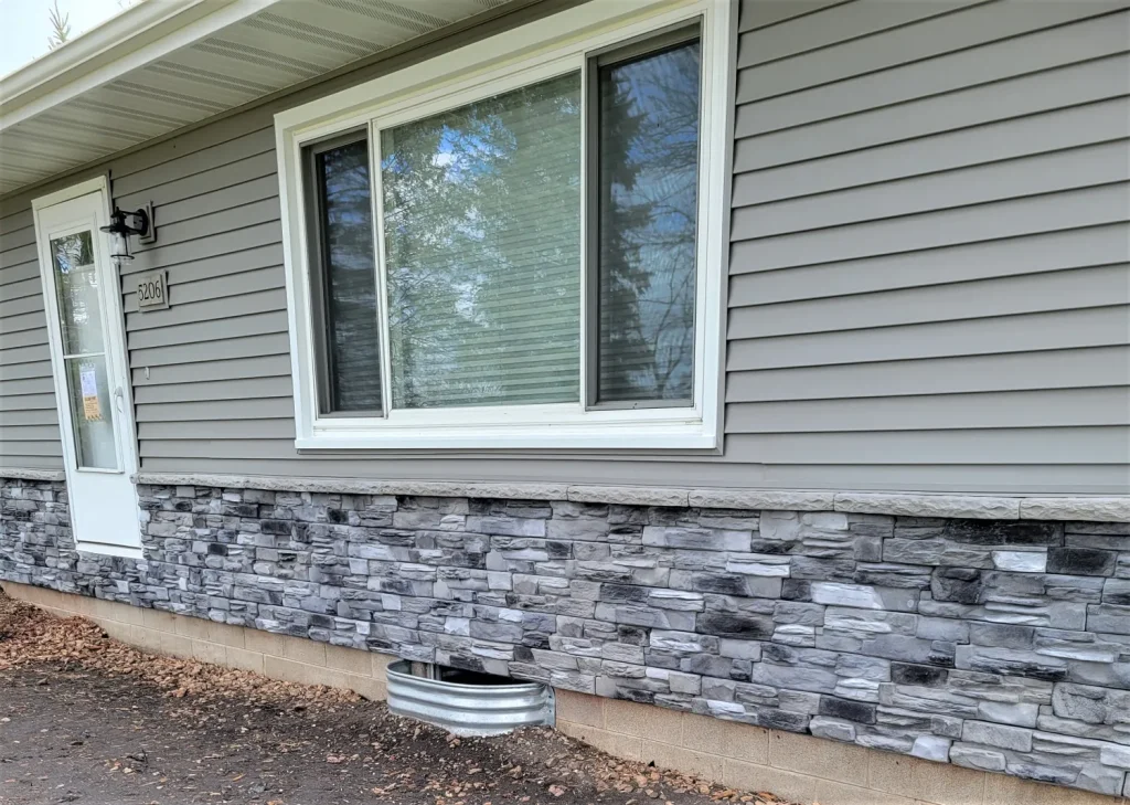 A house exterior with gray siding, stone accent, a large window, and a metal window well below.