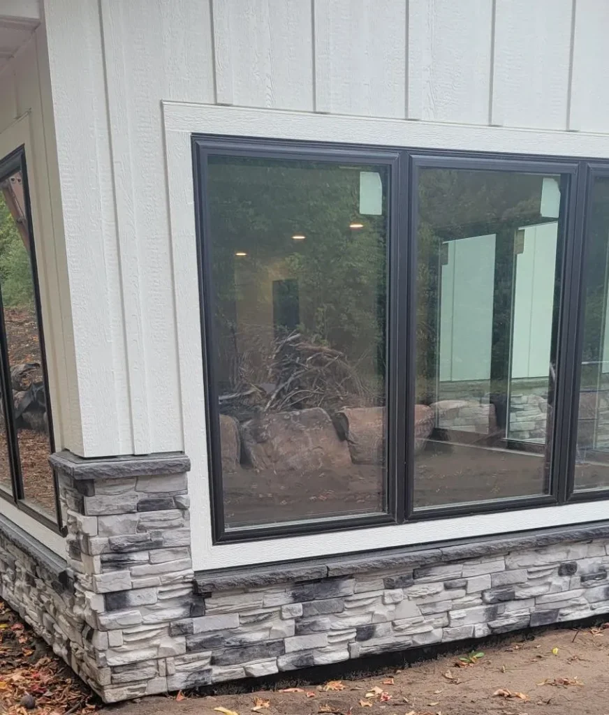 Corner of a house with large windows and gray stone veneer at the base, reflecting outdoor greenery.