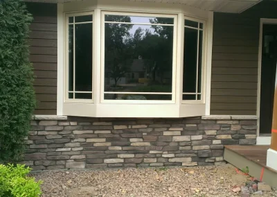 A large bay window with white trim over a stone facade, next to brown siding and a porch pillar.