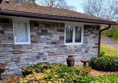 Stone house wall with two windows, a large vase, birdbath, and bushes with fallen autumn leaves.