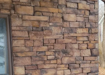 A brick wall with unevenly placed stones next to a window under a roof, with trees visible outside.
