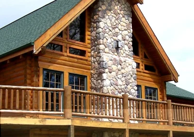 A log cabin with a large stone chimney and wooden balcony, featuring green roof and multiple windows.